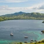 L’île de Padar en Indonésie avec ses eauxbleues foncées, ses traînées turquoise et quelques bateaux dessus, entourée de montagnes vertes.