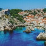 Mer bleue entourant les formations rocheuses vallonnées de Dubrovnik en Croatie avec son port de plaisance, ses maisons blanches aux toits orange, sa forteresse et ses murailles.