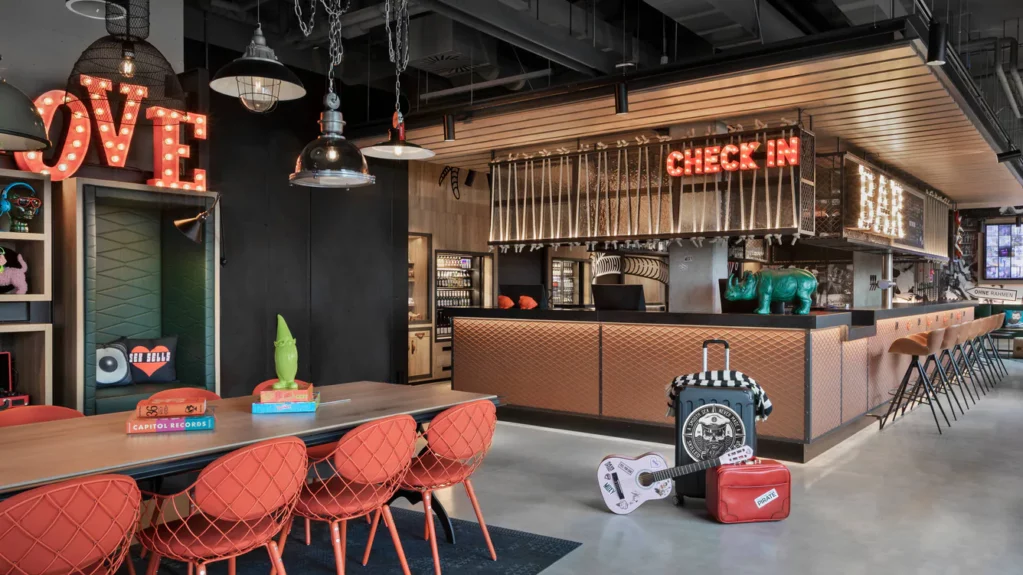 Bronze-toned bar and reception of the Moxy Bremen featuring red neon lights and accent decor pieces, gray and black walls and floors, and a table with red chairs