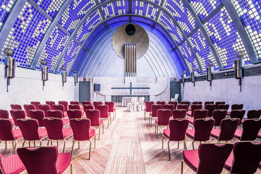 Himmelsaal of the Haus Atlantis with a light wooden floor, rows of red chairs, a blue and clear square mosaic glass roof letting the sun shine through in the whole room