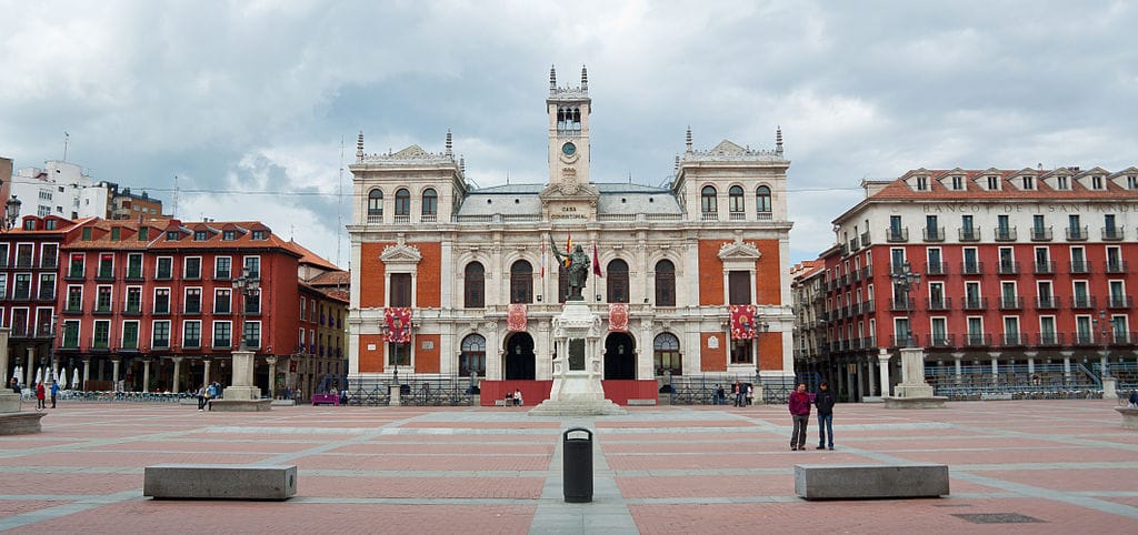 Large mur rouge imposant avec un bâtiment aux détails blancs devant une statue sombre à la la Plaza Mayor au sol rouge.