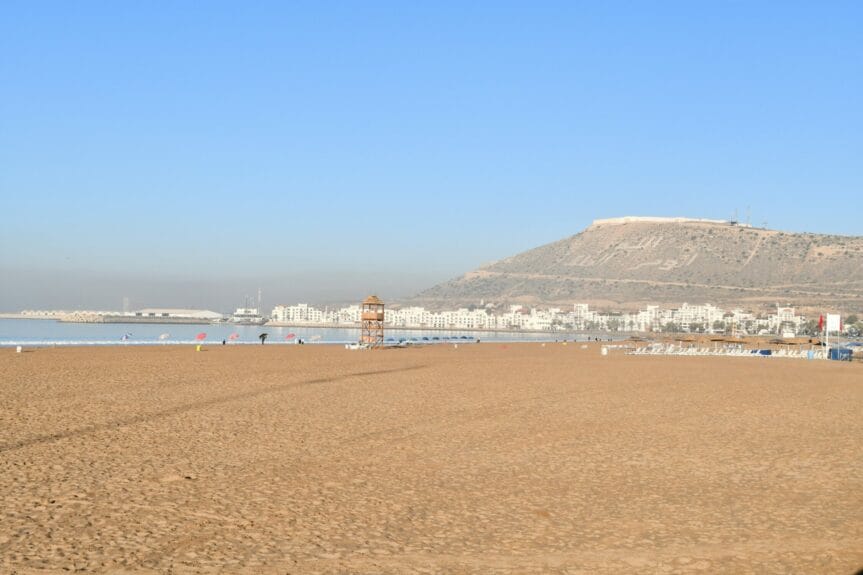 Beds and umbrellas on a beach with mountains and white buildings in the background in Agadir, Morocco.