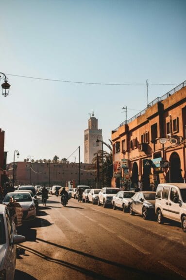 Cars parked on the street in Marrakech, Morocco, near a brown concrete building with a mosque in the background.