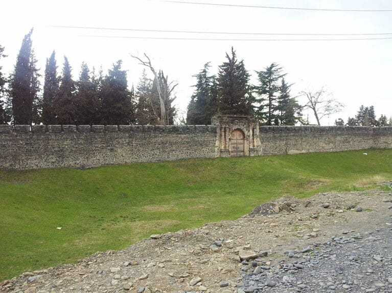 A row of conifer trees behind the ancient brick wall of Azerbaijan’s Zaqatala Fortress with a rounded wooden door in the center standing on a green lawn.