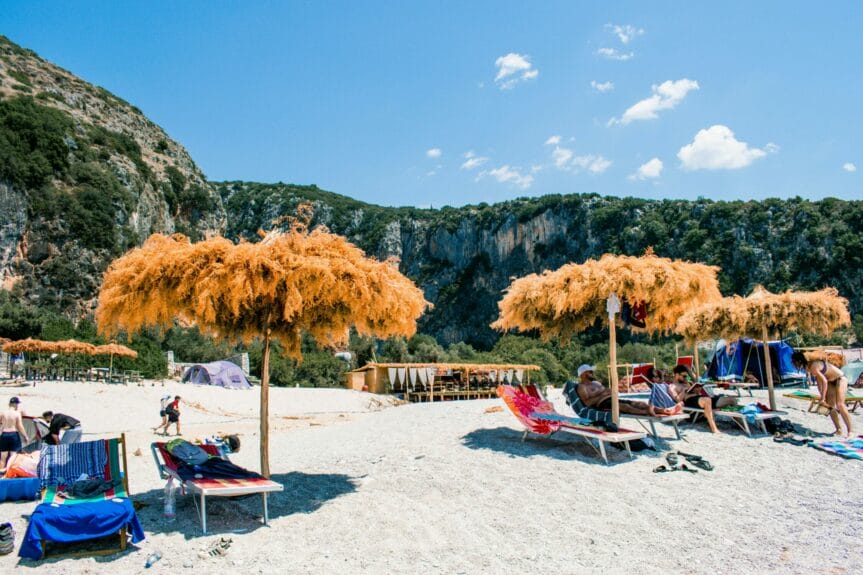  People sitting on chairs with umbrellas made from tree leaves on the shore of Gjipe Beach, Albania with mountains in the background.