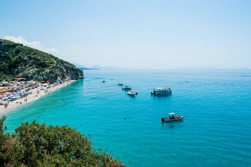 Aerial view of Gjipe Beach, Albania with people, chairs, and umbrellas on the shore and boats on the sea.