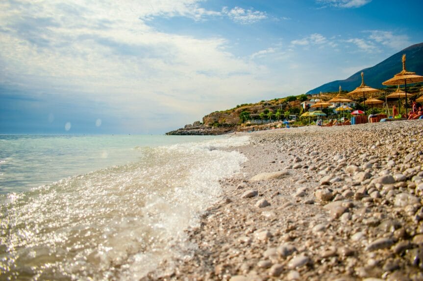 People on the Dhërmi beach, Albania with chairs and umbrellas on the shore and waves crashing on the sand.