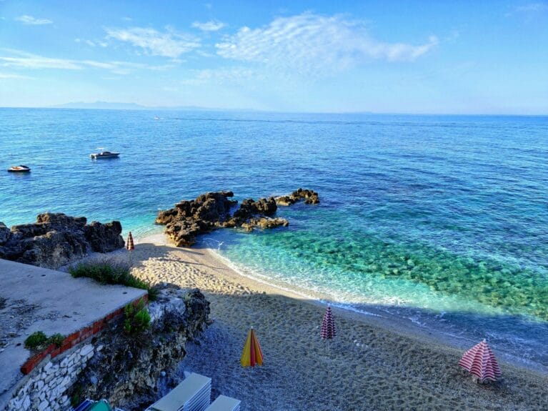 Dhërmi beach, Albania with umbrellas on the sand, boats on the sea, and rock formations.