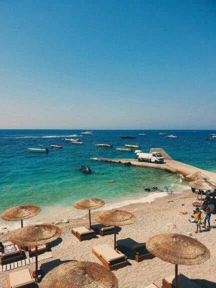 Drymades Beach, Albania with chairs and umbrellas on the shore, boats on the sea, and a white truck on the road by the sea.