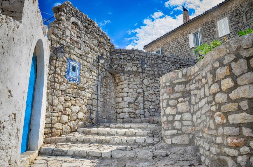 A street in the Himara Castle in Albania with a blue door and walls and floors made of beige stones.