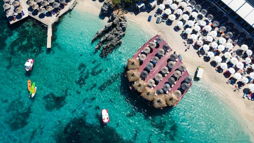 Some boats on the water near a beach filled with people enjoying the sun under shades.