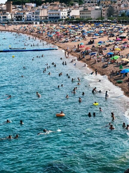 Many people enjoying the beach by the ocean in Tossa de Mar with buildings going up in the mountain in the background on a sunny day. 