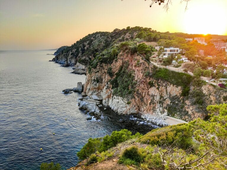 A rocky cliff covered in trees by the ocean with some houses on top at sunset in Tossa de Mar.