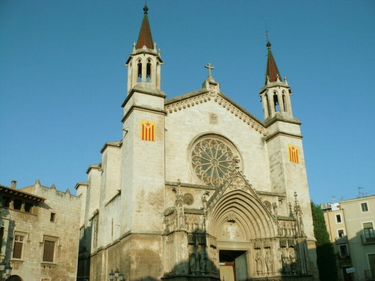 The light brick-walled gothic church of Santa Maria with red and yellow Catalonia flags on each facade of the 2 tours with tall, pointy and colorful roofs in Vilafranca del Penedès.