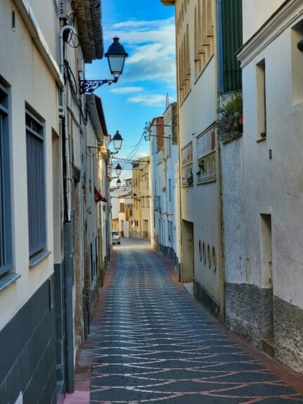 Narrow paved street in Penedès with tall white and yellow buildings on each side on a sunny day.