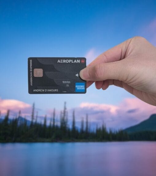 A hand holds an Aeroplan American Express card in front of a peaceful lakeside scene, with trees and mountains under a soft, pastel-colored sky.