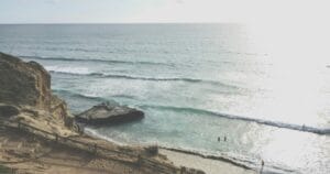 A coastal scene in San Diego, United States, featuring a sandy trail along a cliffside overlooking calm ocean waves, with a few people wading in the shallow waters below.