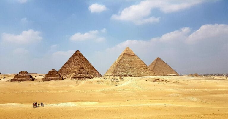 People on the sand near a car on a sunny day, in front of the Pyramids Gardens, Al Haram, in Egypt