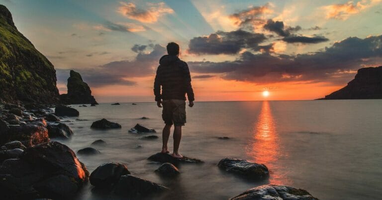 A person stands on rocky shores, gazing at a breathtaking sunset over a calm sea, with vibrant colors reflecting on the water.