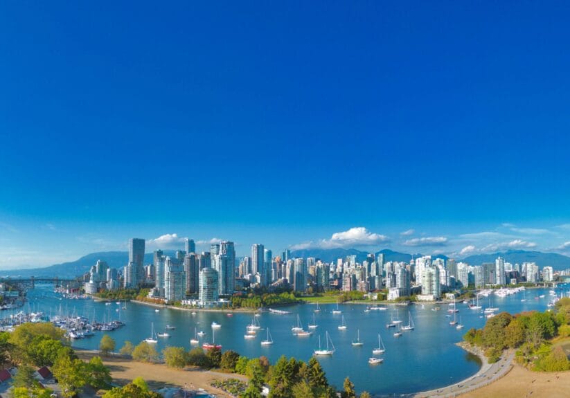 Vue panoramique du centre-ville de Vancouver avec ses gratte-ciels modernes, des voiliers sur False Creek et les montagnes en arrière-plan sous un ciel bleu dégagé.